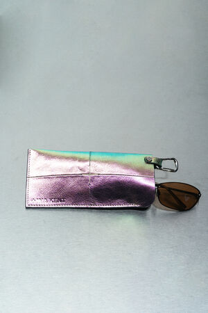 A metallic iridescent glasses case by INA KENT with a silver clasp holds a pair of brown-lensed sunglasses, resting on a light gray surface.