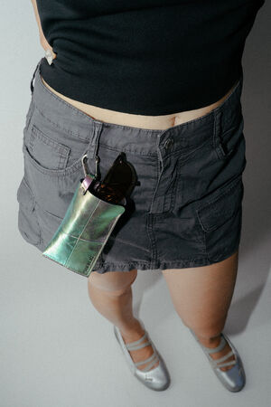 Eine Person, die ein schwarzes Oberteil, einen grauen Cargo-Rock und silberne Schuhe trägt; an der Gürtelschlaufe des Rocks ist eine metallische INA KENT-Tasche befestigt, in der eine Sonnenbrille steckt.