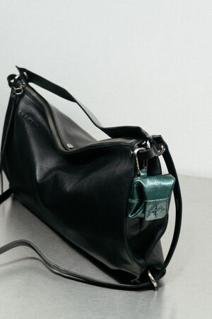 An INA KENT black leather shoulder bag with a silver zipper and a small metallic green pouch attached to its strap, placed on a metallic surface against a plain light background.
