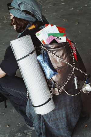 Person wearing a headscarf and black clothes with an INA KENT backpack, holding a rolled yoga mat, books, a blue pouch, a red bandana, metal cup, and beaded chain accessories.